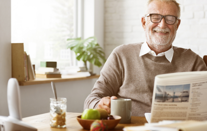 Happy senior man sitting at table reading newspaper while having cup of tea at senior community.