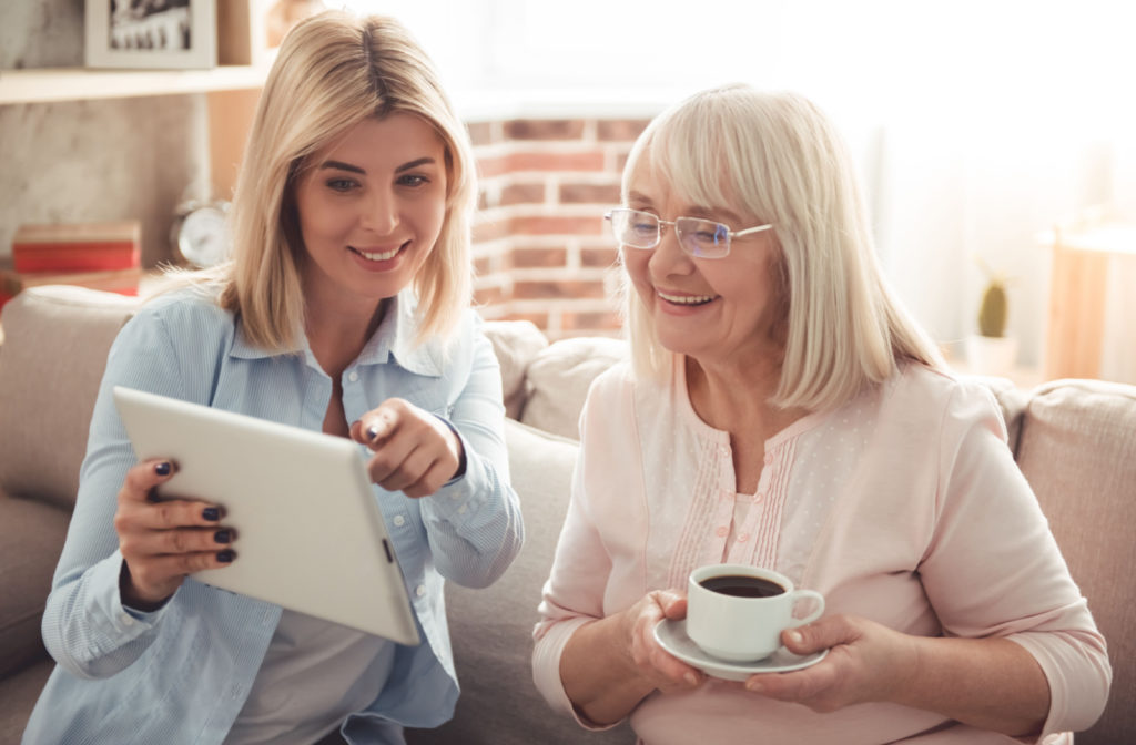 An adult daughter is showing her senior mom a senior living facility online on her tablet while drinking coffee and sitting on a couch at home.