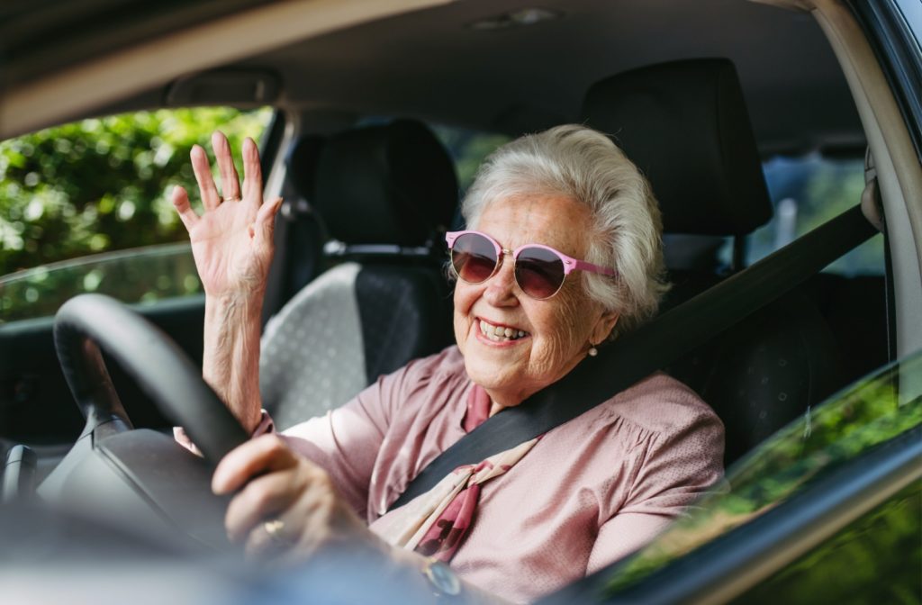 A happy senior in pink sunglasses driving with their right hand raised to wave.