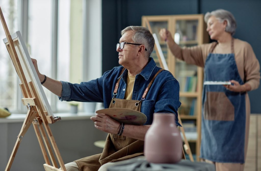 An elderly person and a friend both painting, engaged in their artwork. The scene captures a relaxed, creative moment in a cozy indoor space.