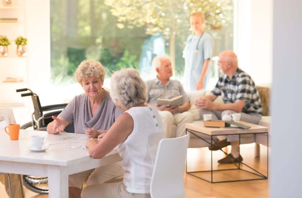 Several older adults sit spread out over a few tables and couches in a shared space of an assisted living community.